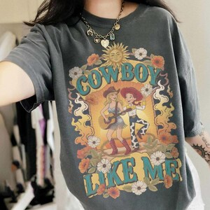 May include: A charcoal gray t-shirt featuring a graphic print with the text "COWBOY LIKE ME" in teal. The design includes a sun, flowers, and two figures, one playing a guitar and the other dancing, surrounded by orange and yellow flame-like shapes.