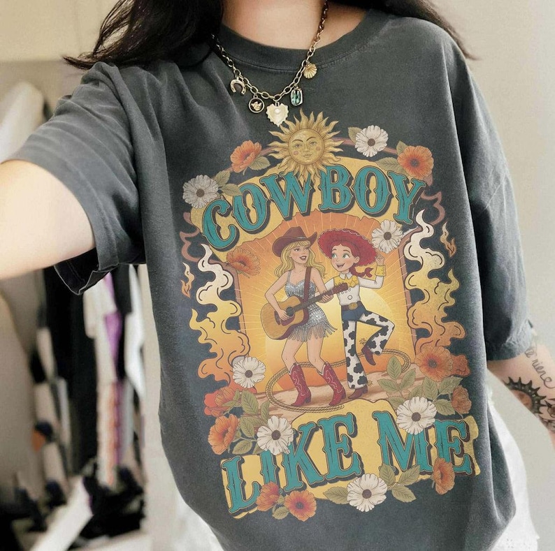 May include: A charcoal gray t-shirt featuring a graphic print with the text "COWBOY LIKE ME" in teal. The design includes a sun, flowers, and two figures, one playing a guitar and the other dancing, surrounded by orange and yellow flame-like shapes.