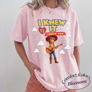 May include: A light pink short-sleeve t-shirt featuring a graphic of a cartoon cowgirl playing an acoustic guitar. The text reads I KNEW IT and I KNEW YOU, surrounded by white clouds, yellow stars, and musical notes. The design includes a red heart.