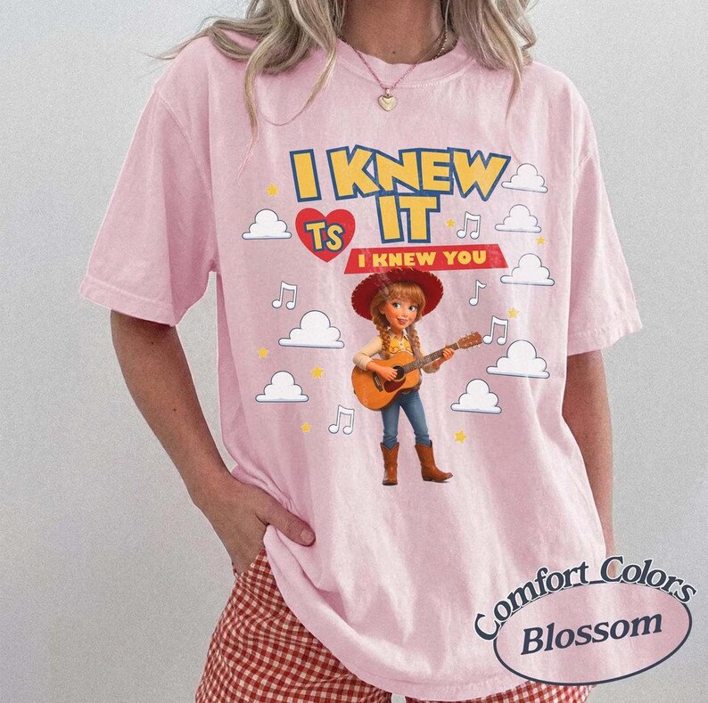May include: A light pink short-sleeve t-shirt featuring a graphic of a cartoon cowgirl playing an acoustic guitar. The text reads I KNEW IT and I KNEW YOU, surrounded by white clouds, yellow stars, and musical notes. The design includes a red heart.