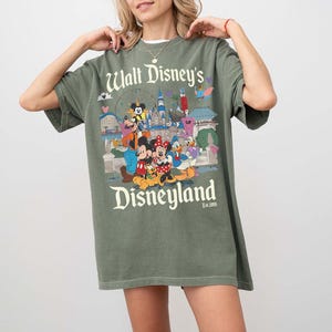 Puede incluir: Camiseta verde oliva con un gr&aacute;fico colorido de personajes de Disney y lugares emblem&aacute;ticos de Disneyland. El texto "Walt Disney's Disneyland" est&aacute; impreso arriba y abajo del gr&aacute;fico. La camiseta tiene mangas cortas y un corte holgado.