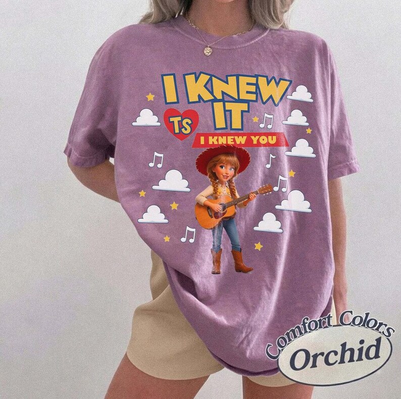 May include: A mauve short-sleeve t-shirt featuring a graphic of a cowgirl playing a guitar. The text reads I KNEW IT and I KNEW YOU, surrounded by white cloud shapes, yellow stars, and musical notes. The shirt has a crew neckline and a relaxed fit.