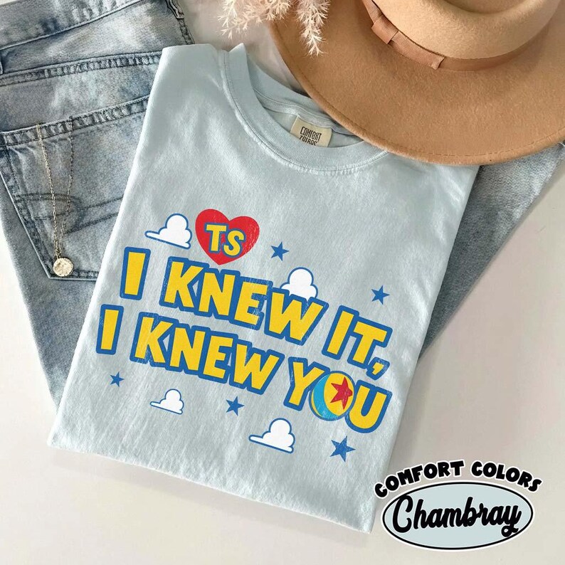 Puede incluir: Una camiseta azul claro con el texto I KNEW IT, I KNEW YOU en amarillo con bordes azules. El dise&ntilde;o incluye un coraz&oacute;n rojo con las letras TS, peque&ntilde;as nubes blancas, estrellas azules y un gr&aacute;fico de pelota colorida con una estrella roja.