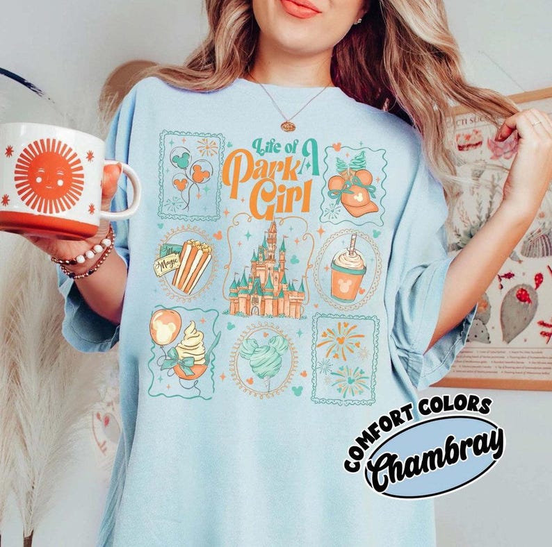 May include: Light blue t-shirt with a colorful graphic design featuring the text "Life of a Park Girl" and various theme park icons. A white mug with a sun design is held in the left hand. The shirt is labeled "Comfort Colors Chambray."