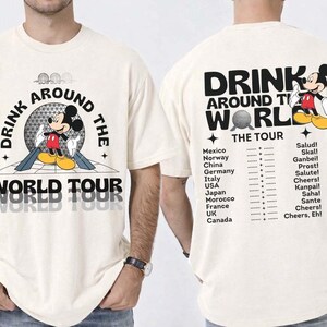 Pode incluir: Camiseta cor marfim com Mickey Mouse e a frase "DRINK AROUND THE WORLD TOUR". A parte de trás mostra países e frases como "Salud!" e "Cheers!" em preto. Uma camiseta divertida e casual.