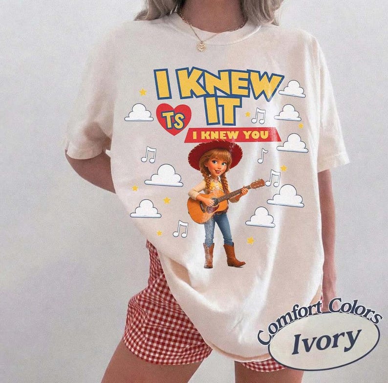 May include: An ivory t-shirt featuring a graphic of a cowgirl character playing an acoustic guitar. The text reads I KNEW IT and I KNEW YOU, accompanied by a red heart, musical notes, and white cloud illustrations scattered across the front.