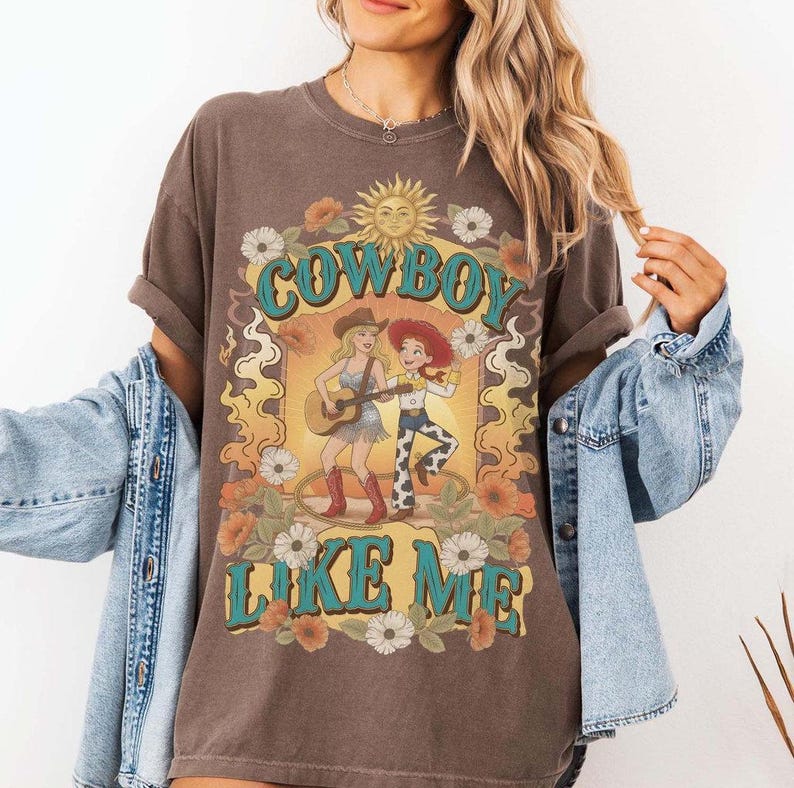 May include: A brown short-sleeve t-shirt featuring a graphic print with the text COWBOY LIKE ME. The design includes two illustrated characters, one playing a guitar, surrounded by a sun, flowers, and flames in yellow, orange, and teal colors.