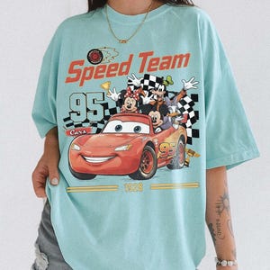 May include: Light blue t-shirt featuring a graphic of Lightning McQueen from the Cars movie with the text "Speed Team 95 Cars 1928". The graphic also includes Mickey Mouse, Minnie Mouse, Goofy, and Donald Duck.