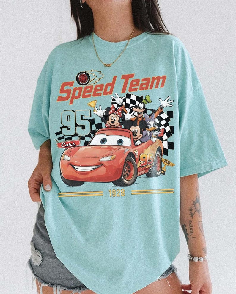 May include: Light blue t-shirt featuring a graphic of Lightning McQueen from the Cars movie with the text "Speed Team 95 Cars 1928". The graphic also includes Mickey Mouse, Minnie Mouse, Goofy, and Donald Duck.