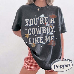 以下が含まれることがあります： 「YOU'RE A COWBOY LIKE ME」という文字がかすれた白のフォントでプリントされた、チャコールグレーの半袖Tシャツ。ロケット、カウボーイハット、保安官の星、ボール、カウボーイブーツの小さなグラフィックイラストがデザインされています。