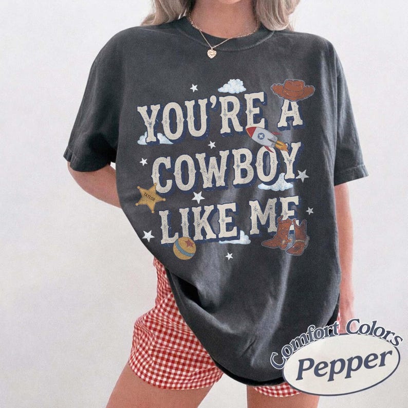 以下が含まれることがあります： 「YOU'RE A COWBOY LIKE ME」という文字がかすれた白のフォントでプリントされた、チャコールグレーの半袖Tシャツ。ロケット、カウボーイハット、保安官の星、ボール、カウボーイブーツの小さなグラフィックイラストがデザインされています。