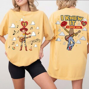 Puede incluir: Una camiseta amarilla con un gr&aacute;fico de una vaquera tocando la guitarra en la parte delantera, rodeada de nubes, notas musicales y un coraz&oacute;n con las letras TS. La parte trasera muestra el texto I KNEW IT I KNEW YOU con un personaje.