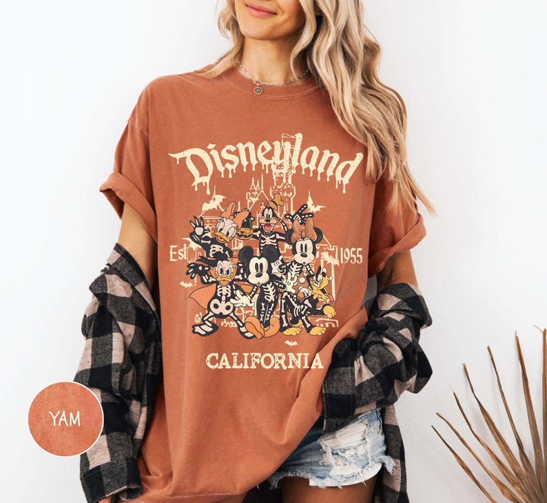 May include: A rust-colored t-shirt featuring a graphic of Disney characters in skeleton costumes. The text "Disneyland" is above the graphic, with "Est 1955" and "California" below. The shirt has rolled-up sleeves.