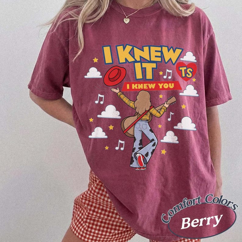 May include: A berry-colored short-sleeve t-shirt featuring a graphic print with the text I KNEW IT and I KNEW YOU. The design includes a person playing an acoustic guitar, a red hat, a red heart with TS, musical notes, stars, and white clouds.