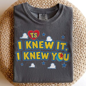Puede incluir: Una camiseta gris carb&oacute;n de cuello redondo con un estampado gr&aacute;fico que incluye el texto "TS" dentro de un coraz&oacute;n rojo, seguido del texto amarillo "I KNEW IT, I KNEW YOU" con una pelota azul y amarilla con una estrella roja.