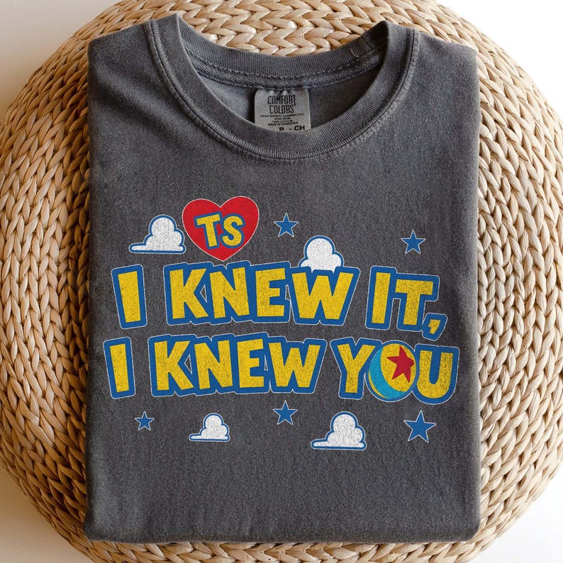 Puede incluir: Una camiseta gris carb&oacute;n de cuello redondo con un estampado gr&aacute;fico que incluye el texto "TS" dentro de un coraz&oacute;n rojo, seguido del texto amarillo "I KNEW IT, I KNEW YOU" con una pelota azul y amarilla con una estrella roja.