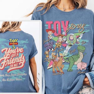 Pode incluir: Uma t-shirt azul com gr&aacute;ficos de Toy Story. A frente mostra as personagens Woody, Buzz Lightyear, Jessie, Rex, Hamm, Bullseye e Slinky Dog com o texto "TOY story". As costas apresentam o texto "TOY STORY 5 You've Got A Friends In Me".