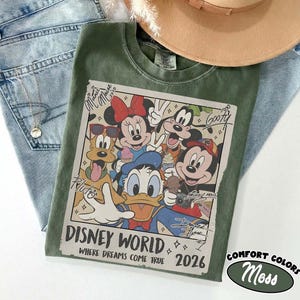 May include: A green Comfort Colors t-shirt featuring a graphic of Disney characters, including Mickey Mouse, Minnie Mouse, Donald Duck, and Goofy. The graphic reads "Disney World Where Dreams Come True 2026". The shirt is paired with blue jeans and a tan hat.