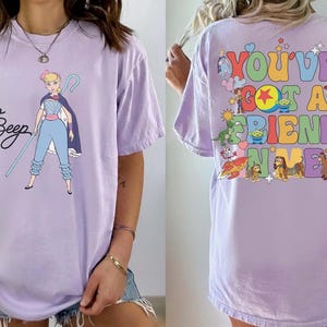 May include: A light purple t-shirt featuring a graphic of Bo Peep on the front with her name in black script. The back displays the colorful text "YOU'VE GOT A FRIEND IN ME" surrounded by various animated character illustrations and small star icons.