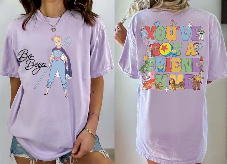 May include: A light purple t-shirt featuring a graphic of Bo Peep on the front with her name in black script. The back displays the colorful text "YOU'VE GOT A FRIEND IN ME" surrounded by various animated character illustrations and small star icons.
