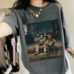 Puede incluir: Camiseta gris oscuro con un gr&aacute;fico de Buzz Lightyear y Woody de Toy Story sentados en una acera. Buzz est&aacute; bebiendo de una botella. La imagen tiene un aspecto vintage y desgastado.