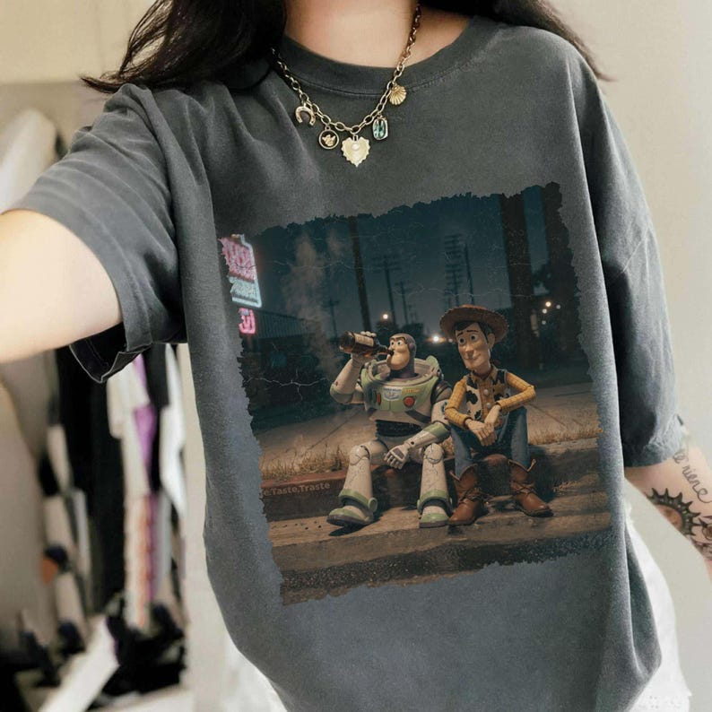 Puede incluir: Camiseta gris oscuro con un gr&aacute;fico de Buzz Lightyear y Woody de Toy Story sentados en una acera. Buzz est&aacute; bebiendo de una botella. La imagen tiene un aspecto vintage y desgastado.