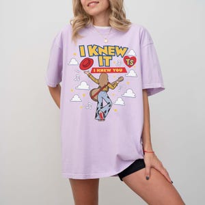 May include: A light purple t-shirt featuring a graphic print of a person playing a guitar, wearing a yellow shirt and cow-print pants. The text reads I KNEW IT I KNEW YOU TS, accompanied by a red hat, a red heart, musical notes, and white clouds.