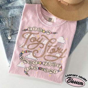 May include: A light pink t-shirt featuring a graphic design with the text "ITS A Tay Tay BABY JUST YES SAY" written in a rope-style font. The design includes colorful beads, stars, musical notes, and white clouds on a soft cotton fabric.