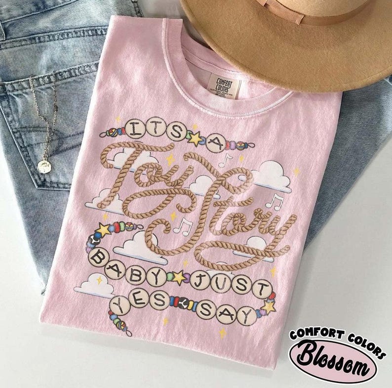 May include: A light pink t-shirt featuring a graphic design with the text "ITS A Tay Tay BABY JUST YES SAY" written in a rope-style font. The design includes colorful beads, stars, musical notes, and white clouds on a soft cotton fabric.