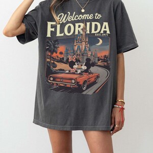 May include: Dark gray t-shirt with a graphic that says "Welcome to Florida" and "Est. 1971." The graphic features Mickey and Minnie Mouse in a red car in front of a castle and palm trees.