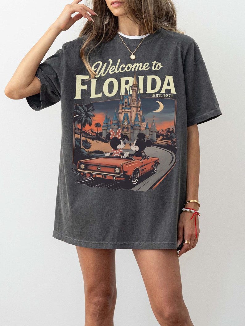 May include: Dark gray t-shirt with a graphic that says "Welcome to Florida" and "Est. 1971." The graphic features Mickey and Minnie Mouse in a red car in front of a castle and palm trees.