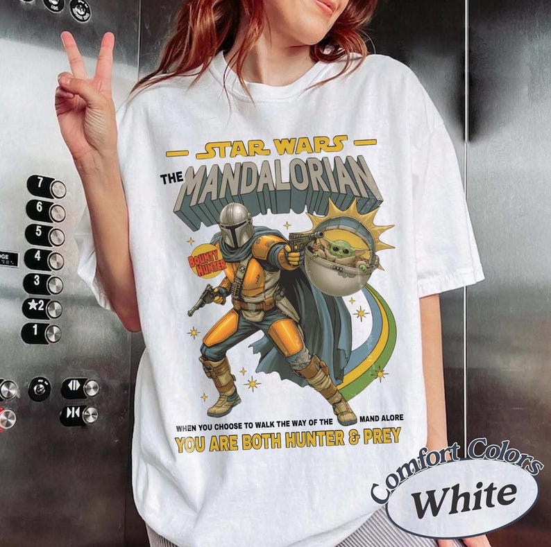 May include: A white t-shirt featuring a graphic print of The Mandalorian in yellow and grey armor holding a blaster, with Grogu in a floating pod. The text reads Star Wars The Mandalorian, Bounty Hunter, and You Are Both Hunter & Prey.