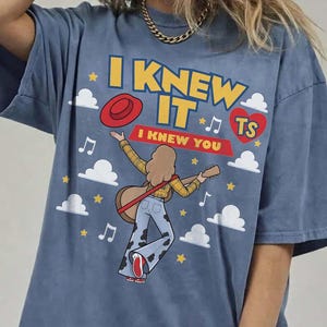 May include: A blue t-shirt featuring a graphic print of a person with long hair playing a guitar, wearing a yellow plaid shirt and cow-print bell-bottom pants. The text reads I KNEW IT and I KNEW YOU, accompanied by a red hat, heart, clouds, and stars.