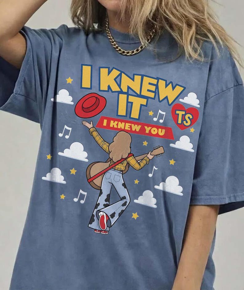 May include: A blue t-shirt featuring a graphic print of a person with long hair playing a guitar, wearing a yellow plaid shirt and cow-print bell-bottom pants. The text reads I KNEW IT and I KNEW YOU, accompanied by a red hat, heart, clouds, and stars.