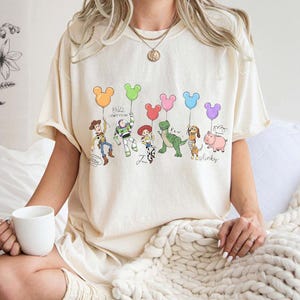 Puede incluir: Camiseta color crema con una ilustraci&oacute;n de dibujos animados de personajes de una pel&iacute;cula animada popular. Los personajes sostienen globos con forma de Mickey Mouse en varios colores. El texto incluye nombres de personajes y "Buzz Lightyear".