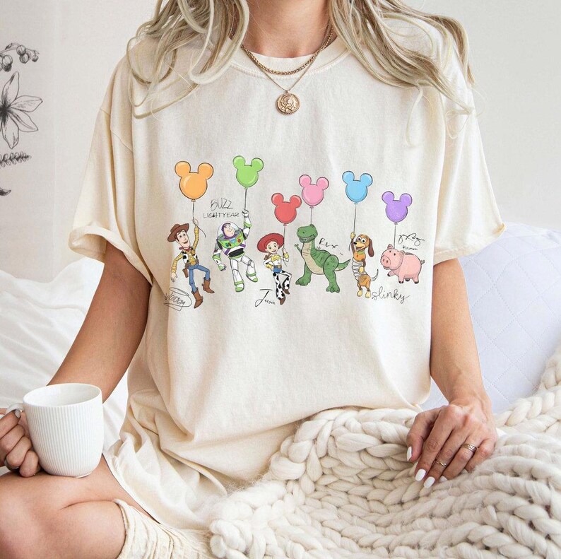 Puede incluir: Camiseta color crema con una ilustraci&oacute;n de dibujos animados de personajes de una pel&iacute;cula animada popular. Los personajes sostienen globos con forma de Mickey Mouse en varios colores. El texto incluye nombres de personajes y "Buzz Lightyear".
