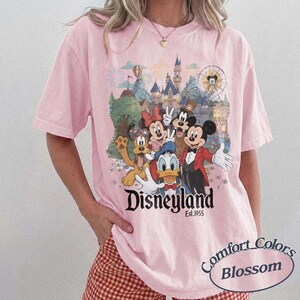 Pu&ograve; includere: Una t-shirt rosa chiaro con una stampa grafica di Topolino, Minnie, Paperino, Pippo e Pluto davanti a un castello. Il testo "Disneyland Est. 1955" &egrave; stampato sotto i personaggi. La t-shirt ha maniche corte e un girocollo.