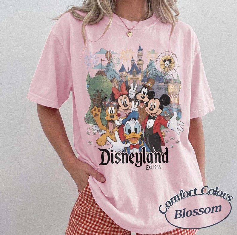 Pu&ograve; includere: Una t-shirt rosa chiaro con una stampa grafica di Topolino, Minnie, Paperino, Pippo e Pluto davanti a un castello. Il testo "Disneyland Est. 1955" &egrave; stampato sotto i personaggi. La t-shirt ha maniche corte e un girocollo.