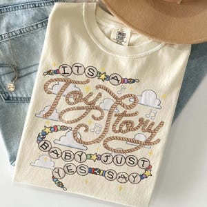 May include: A cream-colored t-shirt featuring a graphic design with the text "ITS A Toy Story BABY JUST YES SAY" written in rope-style lettering and beaded circles. The design includes small clouds, musical notes, and stars in various colors.