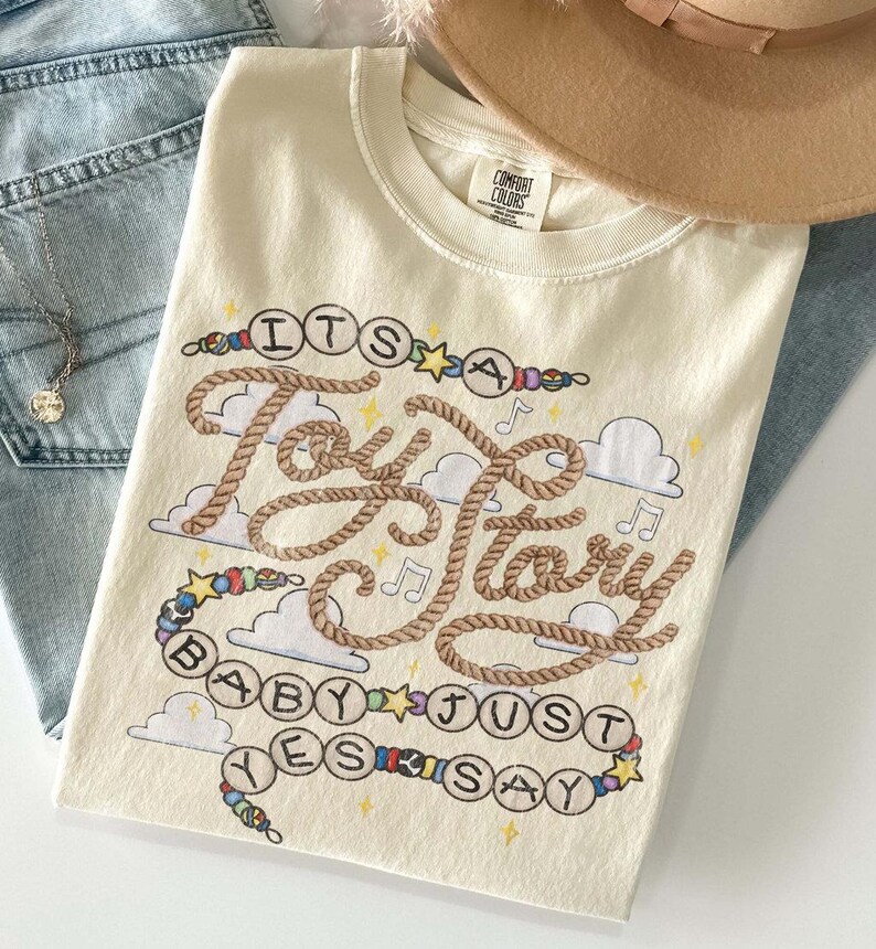 May include: A cream-colored t-shirt featuring a graphic design with the text "ITS A Toy Story BABY JUST YES SAY" written in rope-style lettering and beaded circles. The design includes small clouds, musical notes, and stars in various colors.