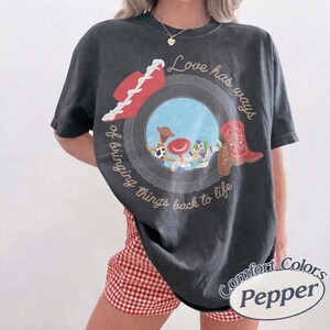 Może przedstawiać: Ciemnoszary t-shirt z kr&oacute;tkim rękawem z okrągłą grafiką przedstawiającą niebieskie niebo, postacie z kresk&oacute;wek, czerwony kowbojski kapelusz i czerwony but. Napis Love has ways of bringing things back to life jest wykonany kursywą na koszulce.