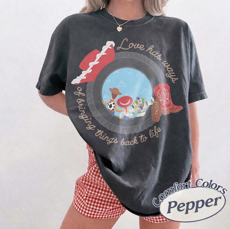 Może przedstawiać: Ciemnoszary t-shirt z kr&oacute;tkim rękawem z okrągłą grafiką przedstawiającą niebieskie niebo, postacie z kresk&oacute;wek, czerwony kowbojski kapelusz i czerwony but. Napis Love has ways of bringing things back to life jest wykonany kursywą na koszulce.