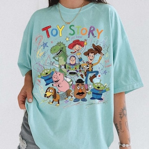 May include: A light blue t-shirt featuring a colorful graphic of characters from the animated film "Toy Story." The design includes the words "Toy Story" in a playful, multi-colored font, along with images of Woody, Buzz Lightyear, and other characters.
