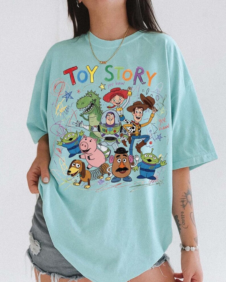 May include: A light blue t-shirt featuring a colorful graphic of characters from the animated film "Toy Story." The design includes the words "Toy Story" in a playful, multi-colored font, along with images of Woody, Buzz Lightyear, and other characters.