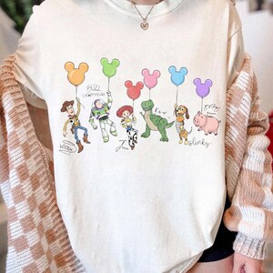 Puede incluir: Sudadera color crema con un gr&aacute;fico colorido de personajes de Toy Story sosteniendo globos con forma de Mickey Mouse. Los personajes incluyen a Woody, Buzz Lightyear, Jessie, Rex, Slinky Dog y Hamm. El texto incluye los nombres de los personajes y "Buzz Lightyear".
