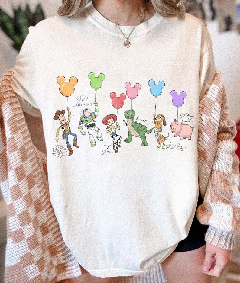 Puede incluir: Sudadera color crema con un gr&aacute;fico colorido de personajes de Toy Story sosteniendo globos con forma de Mickey Mouse. Los personajes incluyen a Woody, Buzz Lightyear, Jessie, Rex, Slinky Dog y Hamm. El texto incluye los nombres de los personajes y "Buzz Lightyear".
