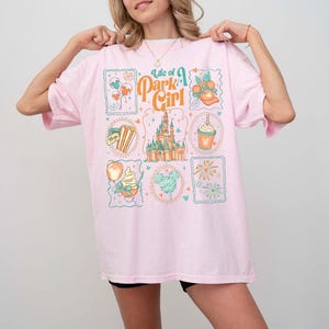 May include: A light pink t-shirt with the text "Life of a Park Girl" and various park-themed graphics. The graphics include a castle, popcorn, ice cream, and fireworks. The shirt is a relaxed fit.