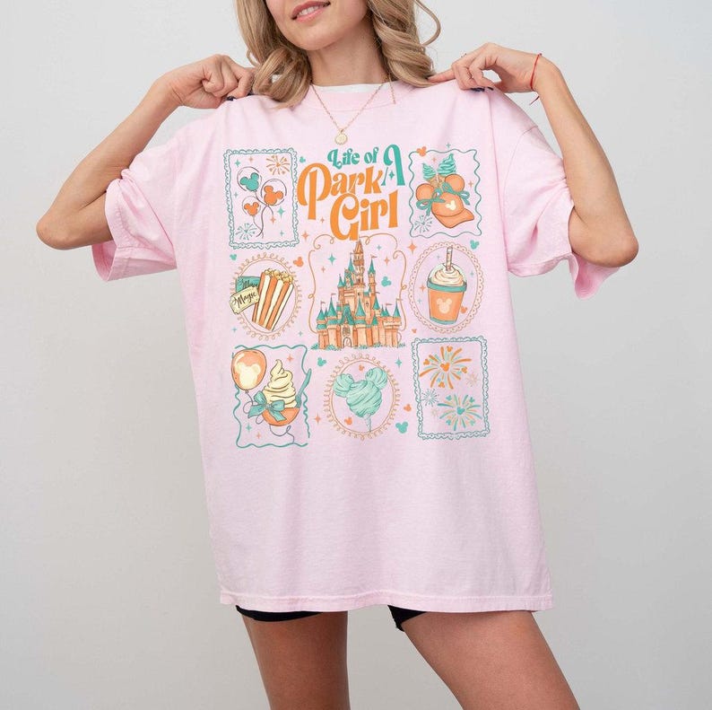 May include: A light pink t-shirt with the text "Life of a Park Girl" and various park-themed graphics. The graphics include a castle, popcorn, ice cream, and fireworks. The shirt is a relaxed fit.