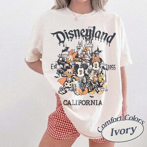 May include: Ivory-colored t-shirt with a Disneyland graphic featuring cartoon characters in skeleton costumes. The design includes the text "Disneyland," "Est. 1955," and "California." The shirt is a Comfort Colors brand.