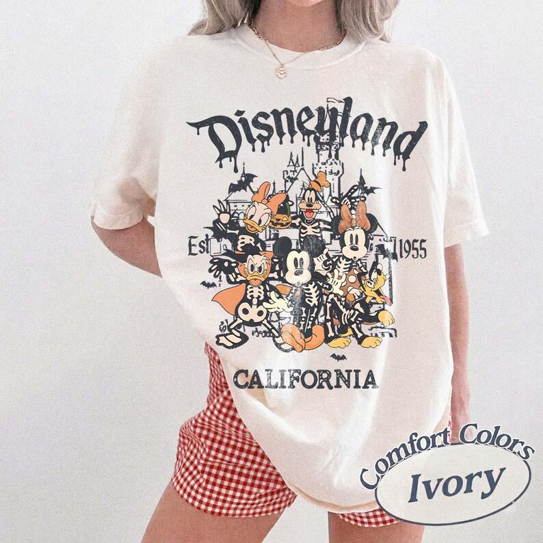 May include: Ivory-colored t-shirt with a Disneyland graphic featuring cartoon characters in skeleton costumes. The design includes the text "Disneyland," "Est. 1955," and "California." The shirt is a Comfort Colors brand.